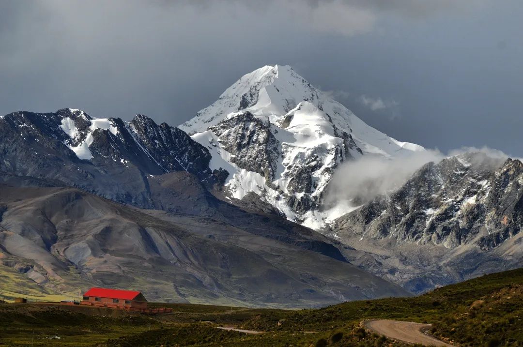 Huayna Potosí seen from the altiplano: its snow face under storm cloud, a small red-roofed refuge at the foot of the slope for scale.